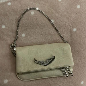 Beige axelväska från Zadig & Voltaire - Snygg beige axelväska i skinn från Zadig & Voltaire med silverdetaljer och kedjerem. Väskan har en dekorativ vingdetalj på framsidan och dragkedja både fram och bak. Perfekt för att ge din outfit en stilren touch.