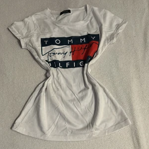 Vit t-shirt från Tommy Hilfiger - Snygg vit t-shirt från Tommy Hilfiger med ett stort tryck i blått och rött på framsidan. Den har en rund halsringning och korta ärmar. Perfekt för en avslappnad stil.