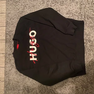 Svart tröja från Hugo - Helt ny utan pris lapp, Storlek S.  Snygg svart tröja från Hugo med deras logga i vitt och rött på framsidan. Tröjan har långa ärmar och ribbade muddar vid ärmslut och nederkant. Perfekt för en stilren look.