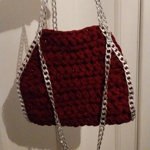 Vinröd virkad handväska med silverdetaljer - Unik vinröd virkad handväska med silverkedjor som handtag och detaljer längs kanterna. Perfekt för att ge en färgklick till din outfit. Väskan har en charmig och handgjord känsla.