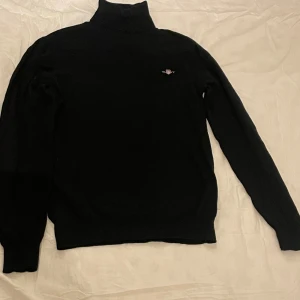 Svart polotröja från Gant - Snygg svart polotröja med en diskret broderad logga på bröstet. Tröjan har långa ärmar och en klassisk polokrage, perfekt för kyligare dagar. Passar både till vardags och mer uppklädda tillfällen.