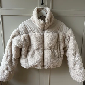 Beige teddyjacka med dragkedja - Mysig beige teddyjacka med dragkedja och hög krage. Perfekt för kyliga dagar med sin fluffiga och varma design. Jackan har en croppad passform och långa ärmar.