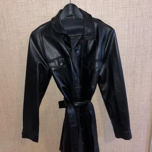 Svart skinnjacka med bälte - Snygg svart skinnjacka med knappar och bälte i midjan. Jackan har långa ärmar och två bröstfickor med lock. Perfekt för en stilren look.