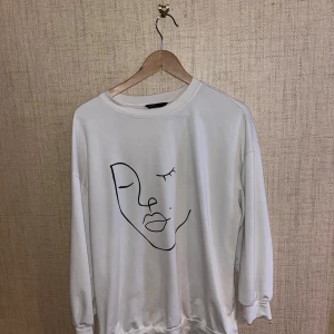 Vit sweatshirt med ansiktsmotiv - Säljer en stilren vit sweatshirt med ett minimalistiskt ansiktsmotiv i svart på framsidan. Tröjan har långa ärmar och en rund halsringning. 
