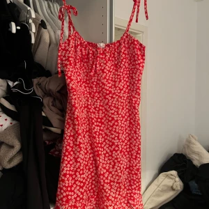 Röd blommig klänning med knytband - Charmig röd klänning med vita blommor och knytband vid axlarna. Perfekt för sommarens varma dagar. Den har en smockad överdel som ger en bekväm passform och en luftig kjol.