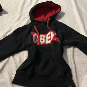 Svart hoodie från Obey - Säljer en svart hoodie från Obey med röd insida i huvan och röda snören. Den har en stor vit och röd logga på framsidan. Perfekt för en avslappnad stil.