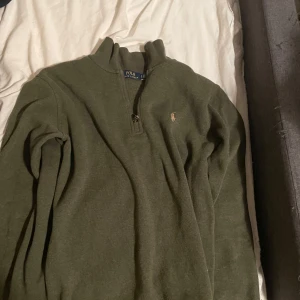 Olivgrön tröja från Polo Ralph Lauren - Säljer en olivgrön tröja från Polo Ralph Lauren med dragkedja vid halsen och broderad logga på bröstet. Tröjan har långa ärmar och en ribbad krage. Perfekt för en stilren och avslappnad look.