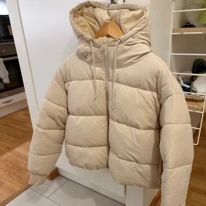 Beige pufferjacka med huva - Säljer en snygg beige pufferjacka med huva. Jackan har en dragkedja framtill och är quiltad för extra värme. Den har en avslappnad passform och långa ärmar.
