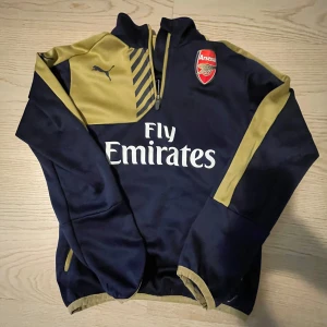 Mörkblå och guldfärgad Arsenal tröja från Puma - Säljer en snygg mörkblå och gul Arsenal tröja från Puma med klubbens emblem på bröstet. Tröjan har en dragkedja vid halsen och långa ärmar. Perfekt för fotbollsfans!