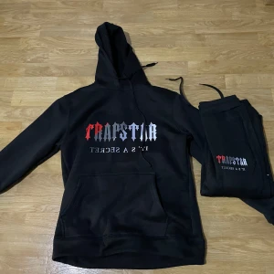 Svart hoodie från Trapstar - Säljer en svart hoodie från Trapstar med tryck i vitt och rött på framsidan. Hoodien har en stor ficka framtill och justerbar huva. Perfekt för en avslappnad stil.