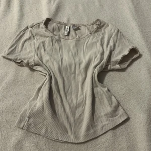 Vit ribbad topp från H&M Divided - Säljer en vit ribbad topp från H&M Divided. Toppen har korta ärmar och en rund halsringning med fina spetsdetaljer. Perfekt för en avslappnad stil.