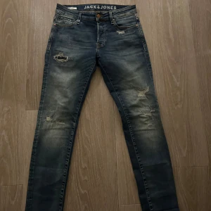 Blå jeans från Jack & Jones - Snygga blå jeans från Jack & Jones med slitna detaljer och en slim passform. De har en klassisk femficksdesign och en läderpatch med logotyp baktill. Perfekta för en avslappnad stil.