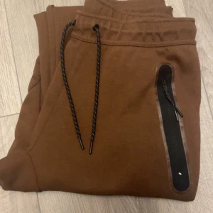 Bruna mjukisbyxor från Nike - Snygga bruna mjukisbyxor från Nike med praktiska dragkedjor på fickorna. Byxorna har en elastisk midja med snörning för bästa passform. Perfekta för en avslappnad stil.