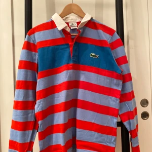 Randig pikétröja från Lacoste - Säljer en färgglad randig pikétröja från Lacoste i rött och blått. Tröjan har långa ärmar och en klassisk krage med knappar. Stor i storlek S.