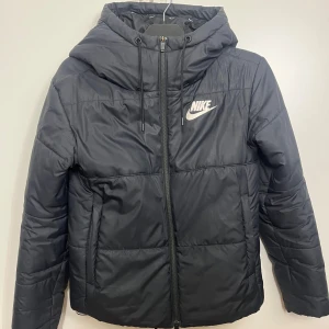 Svart pufferjacka från Nike - Säljer en svart pufferjacka från Nike med huva och dragkedja. Jackan har en stilren design med Nike-logga på bröstet och är perfekt för kyligare dagar. Den är långärmad och har en bekväm passform.