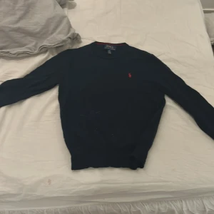 Svart tröja från Ralph Lauren - Säljer en klassisk mörkblå tröja från Ralph Lauren med det ikoniska röda logotypbroderiet på bröstet. Tröjan har långa ärmar och en rund halsringning. Perfekt för en stilren look.