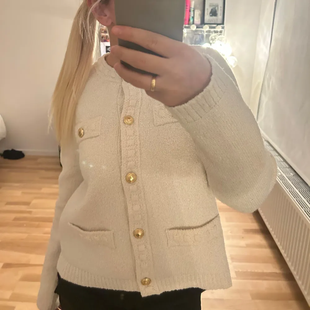 Säljer en stilren vit kofta med eleganta guldknappar. Koftan har långa ärmar och två fickor framtill. Super fin!. Neuletakit & Villapaidat.