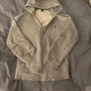 Grå hoodie från Ralph Lauren - Säljer en stilren grå hoodie från Ralph Lauren med dragkedja och klassisk logga på bröstet. Perfekt för en avslappnad look. Hoodien har långa ärmar och en bekväm passform.