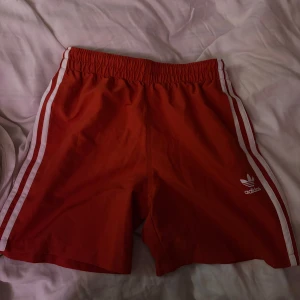Röda shorts från Adidas - Säljer ett par röda balla shorts från Adidas med de klassiska vita ränder längs sidorna. Så snyggt till sommaren med ett vitt linne!!  Elastisk midja och najs material 👌🙌Oanvända