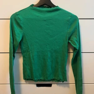 Grön ribbad tröja - Snygg grön ribbad tröja med långa ärmar och volangdetaljer vid hals och nederkant. Perfekt för en stilren look.
