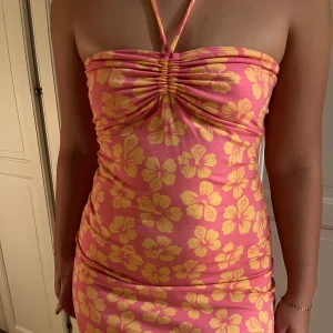 Rosa blommig hawaii klänning - Säljer en söt rosa halter klänning med gula blommor från Hollister. Klänningen är axelbandslös med en halterneck-knytning och har en tight passform. Med en scrunch vid brösten🩷🧡💛🌺