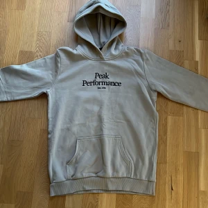 Beige hoodie från Peak Performance - Säljer en snygg beige hoodie från Peak Performance i storlek 170. Den har en klassisk design med svart logga på bröstet och en praktisk magficka. Perfekt för både vardag och chill. Huvan är mjuk och bekväm, perfekt för kyliga dagar. 