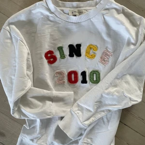 Vit collage tröja från By Malina - Säljer snygg tröja från Malina med färgglada bokstäver som bildar texten 'SINCE 2010'. Tröjan har långa ärmar och är gjord i mjukt material med ribbade muddar vid ärmslut och nederkant. Perfekt för en avslappnad stil! 🌟