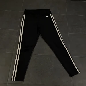 Svarta leggings från Adidas - Säljer ett par svarta leggings från Adidas i storlek M. De har de klassiska tre vita ränderna längs sidorna och är perfekta för träning. Materialet är mjukt och stretchigt, vilket ger en bekväm passform. Perfekta för gymmet eller en avslappnad dag hemma.