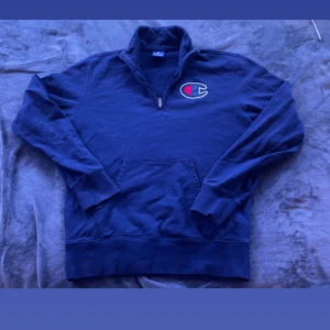 Mörkblå tröja från Champion - Säljer en mörkblå tröja från Champion i bomull strorlek M. Den har en halv dragkedja och en stor ficka fram. Det finns en liten fläck på ärmen, men annars i bra skick. Perfekt för höst och vinter!