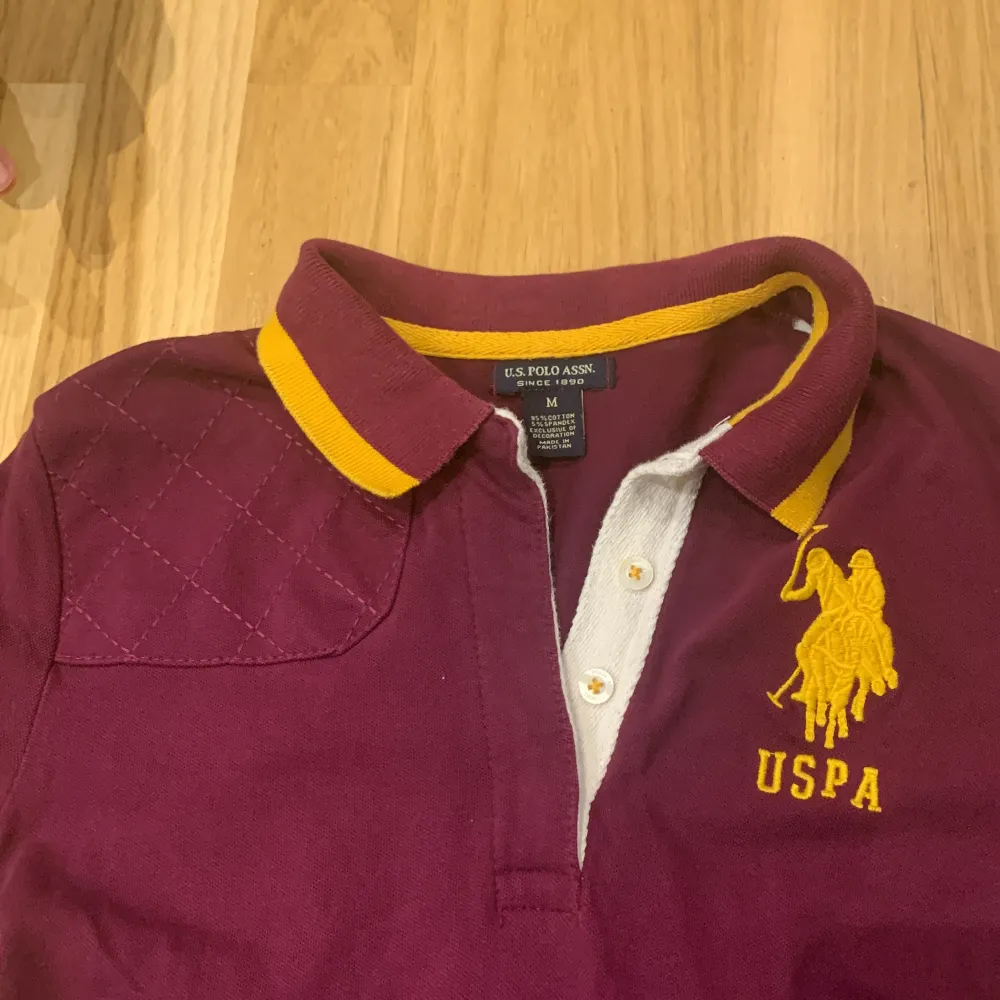 Säljer en snygg vinröd/lila klänning från U.S. Polo Assn. med gul broderad logga på bröstet. Klänningen har en klassisk pikékrage med gula detaljer och knappar framtill. Perfekt för en avslappnad men stilren look. Tillverkad i 95% bomull och 5% spandex för en bekväm passform.. Klänningar.