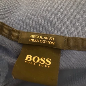 Blå pikétröja från Hugo Boss - Säljer en stilren blå pikétröja från Hugo Boss i regular fit. Tillverkad av mjuk Pima bomull, perfekt för både vardag och lite finare tillfällen. Tröjan har korta ärmar och en klassisk krage med knappar. Passar perfekt till jeans eller chinos!