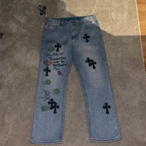 Unika jeans med tryck från Levi's och Chrome Hearts - Säljer ett par unika ljusblå jeans från Levi's i samarbete med Chrome Hearts och CDG. De har coola svarta korsdetaljer och texttryck på framsidan. Perfekta för att sticka ut på festen eller bara ha en chill dag. De är i bra skick och har en straight passform.
