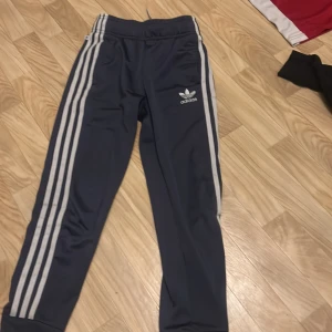 Mörkblå träningsbyxor från Adidas - Säljer ett par mörkblå träningsbyxor från Adidas i storlek 140. Byxorna har de klassiska tre vita ränderna längs sidorna och en elastisk midja för extra komfort. Perfekta för träning eller en avslappnad dag. De är i mycket bra skick och redo för nya äventyr!