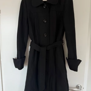 Svart kappa med bälte Lindex - Snygg svart kappa i mycket bra skick. Den har långa ärmar och knappar framtill samt ett bälte i midjan för en elegant look. Perfekt för höst och vinter, både stilren och praktisk. Passar till alla tillfällen!