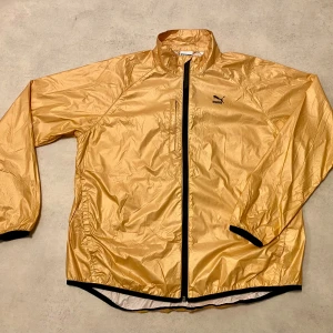 Guldig vindjacka från Puma XL/42 - Säljer en snygg gul eller mer guldfärgad vindjacka från Puma i mycket bra skick. Jackan har en dragkedja framtill och är långärmad med svarta detaljer vid ärmslut och dragkedja. Perfekt för vår och höst när det blåser lite extra. Storleken är XL/42.