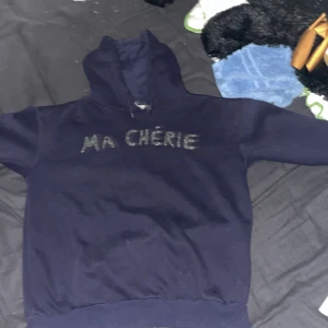 Mörkblå hoodie från Gina Tricot - Säljer en snygg mörkblå hoodie från Gina Tricot med texten 'MA CHÉRIE' i glittriga stenar på bröstet. Perfekt för en casual look eller mysiga dagar hemma. Den har en klassisk passform med huva och ribbade muddar. Passar perfekt till jeans eller leggings!