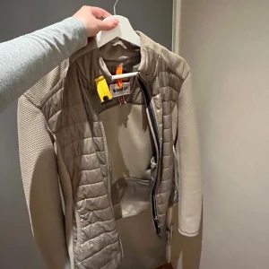 Beige jacka från Parajumpers - Säljer en snygg beige jacka från Parajumpers i mycket bra skick. Jackan har en quiltad design med dragkedja och långa ärmar. Perfekt för höst och vår, och den har en cool detalj med en gul rem vid kragen. Passar både till vardags och lite finare tillfällen.
