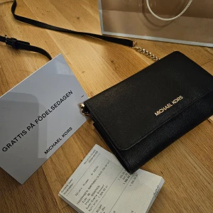 Svart axelväska från Michael Kors - Snygg svart axelväska från Michael Kors i skinn med guldfärgad kedja. Perfekt för både vardag och fest. Väskan har flera fack och en dragkedja för säker förvaring. Stilren design med Michael Kors-logga på framsidan. Mobilförvaring på väskans baksida. 