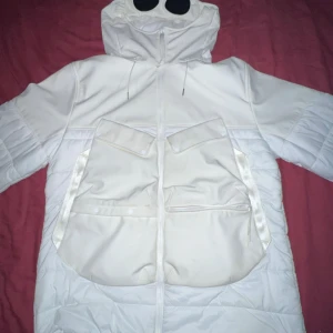 Vit pufferjacka från C.P. Company - Säljer en snygg vit pufferjacka från C.P. Company i storlek M. Jackan har en cool design med huva och stora fickor framtill. Den är gjord av polyester och har en dragkedja. Perfekt för vintern och håller dig varm och stilren! ❄️ har kvitto 