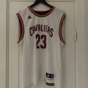 Vit Cavaliers basketlinne från Adidas - Säljer ett snyggt och sportigt basketlinne från Adidas i vitt med Cavaliers tryck och nummer 23. Linnet är ärmlöst och perfekt för basketträning eller som en cool vardagsoutfit. Det är i mycket bra skick och redo för en ny ägare!