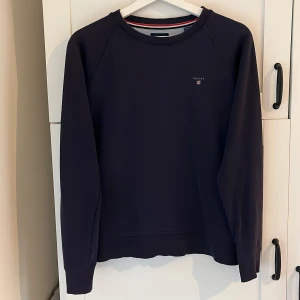 Mörkblå tröja från GANT - Säljer en stilren mörkblå tröja från GANT i storlek 158/164 cm, perfekt för tonåringar. Tröjan har en klassisk design med ribbad krage och ärmar. Den är i bra skick och passar både till vardags och lite finare tillfällen. Superbekväm och lätt att matcha med andra plagg! Nypris 650