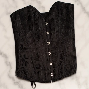 Svart korsett med snörning - Snygg svart korsett med elegant mönster och snörning bak. Den har hakknäppning fram och är perfekt för festliga tillfällen. Materialet är glansigt och ger en lyxig känsla. Passar bra att styla med både kjol och byxor för en chic look. Plagget är aldrig använt då den är för stor för mig. Skickar samma dag som den blir köpt (undantag om det är sent)  