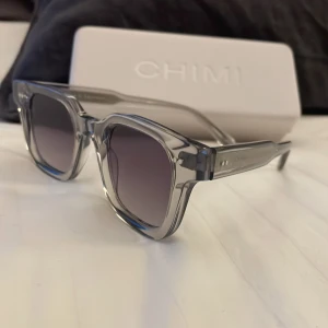  CHIMI 04 i grå - Säljer ett par snygga grå solglasögon från CHIMI i nytt skick. De har en stilren, fyrkantig design med transparenta bågar och mörka linser. Perfekta för både vår och sommar! Kommer med ett skyddande fodral.