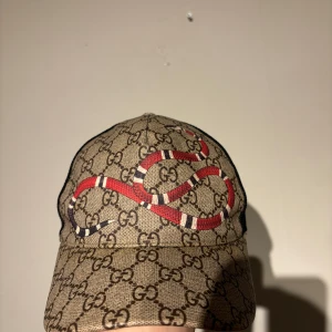 Gucci keps med ormmönster - Säljer en snygg Gucci keps i beige med det klassiska GG-mönstret och en cool broderad orm i rött och vitt. Kepsen har justerbar rem baktill med gröna och röda detaljer. Perfekt för att ge din outfit en lyxig touch!