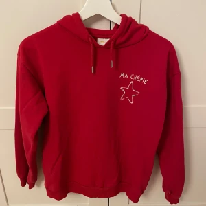 Röd hoodie från Gina Tricot - Säljer en röd hoodie från Gina Tricot med texten 'MA CHÉRIE' och en stjärna på bröstet. Tröjan har en klassisk huva med dragsko och långa ärmar. Perfekt för en avslappnad stil.