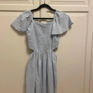 Ljusblå klänning från Kappahl - Charmig ljusblå klänning från Kappahl med offshoulder-design och korta ärmar. Klänningen har en smockad överdel och en luftig kjol, perfekt för varma dagar. Den är tillverkad i ett mjukt och bekvämt material.