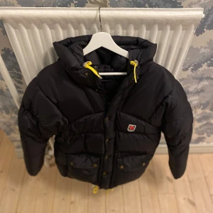 Svart dunjacka från Fjällräven - Säljer en svart dunjacka från Fjällräven med huva och gula detaljer. Jackan har en broderad logotyp på bröstet och stängs med dragkedja och knappar. Perfekt för kalla dagar!