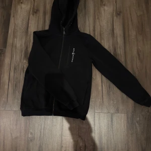 Svart huvtröja från Sail Racing - Snygg svart huvtröja från Sail Racing med dragkedja framtill och en praktisk bröstficka. Perfekt för en avslappnad stil med långärmade ärmar och mjukt material.