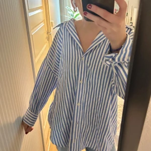 Blå och vit randig skjorta - Säljer en stilren blå och vit randig skjorta med knappar framtill. Skjortan har en avslappnad passform och långa ärmar, perfekt för en casual look. Tillverkad i Marocko.