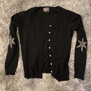 Svart kofta från Culture med stjärndetaljer - Snygg svart kofta från Culture med vita knappar och coola stjärndetaljer på ärmarna. Perfekt för att ge din outfit en unik touch. Långärmad och bekväm passform.